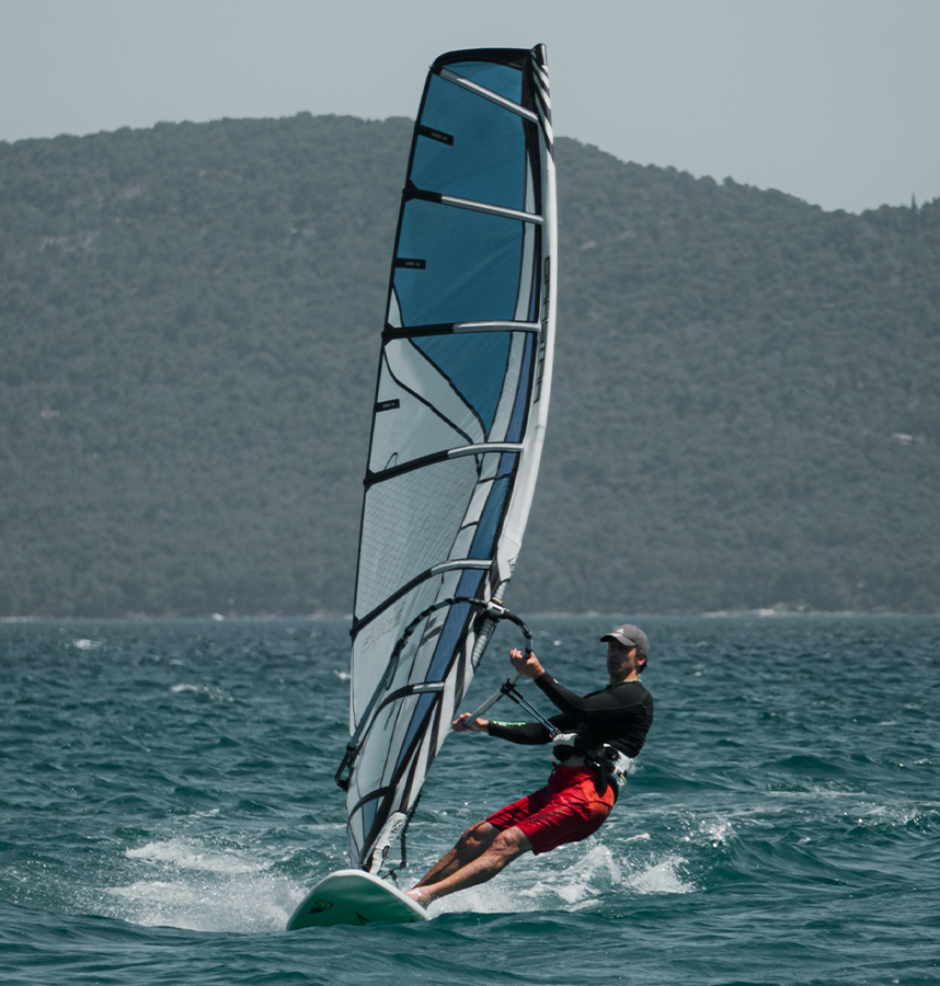 Zdjęcie przedstawia mężczyznę na windsurfingu. W tle jest wzniesienie porośnięte drzewami. 