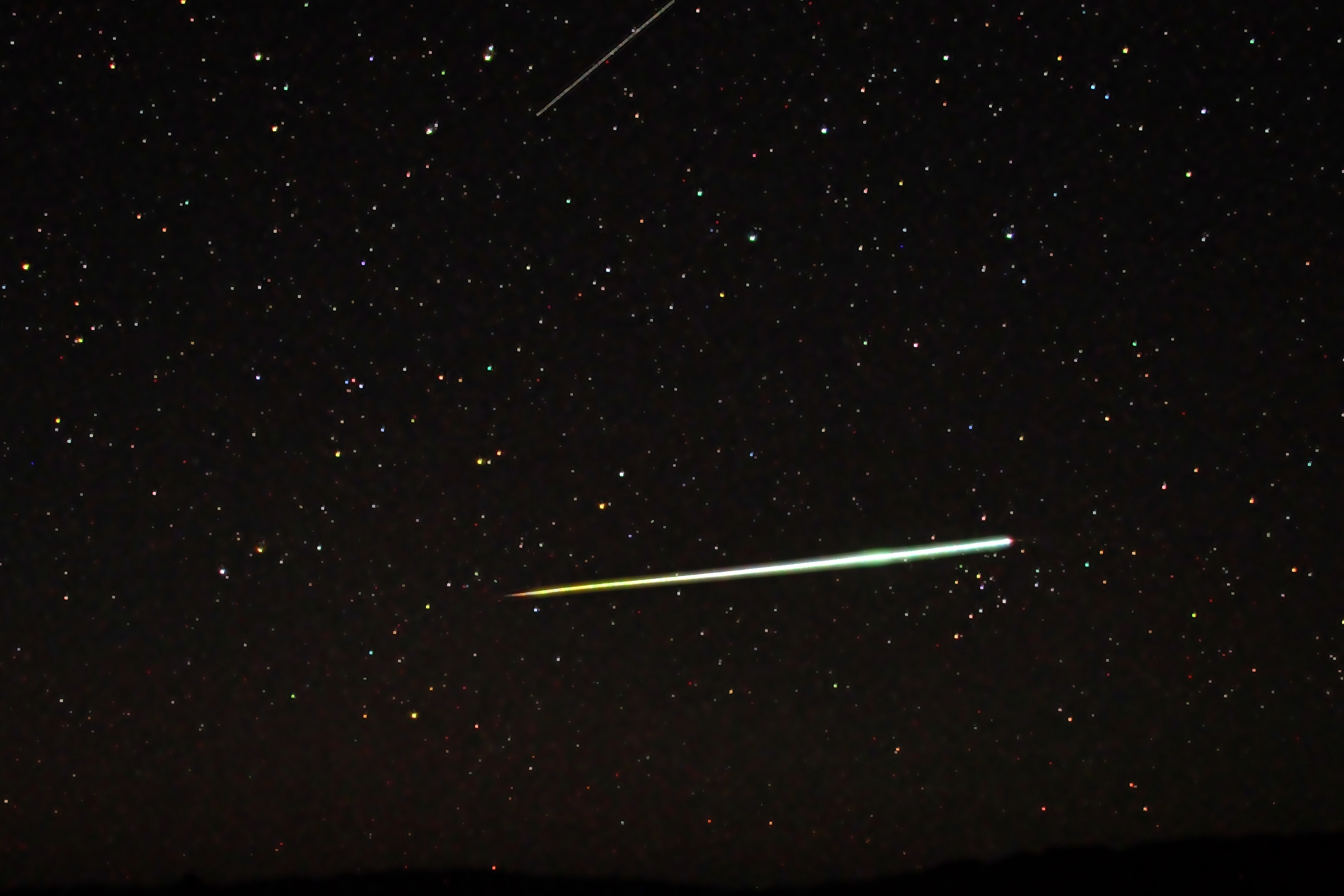 Fotografia prezentuje meteor. Na czarnym niebie widoczne gwiazdy w postaci białych licznych punkty różnej wielkości. Na środku fotografii widoczna biała wąska smuga.