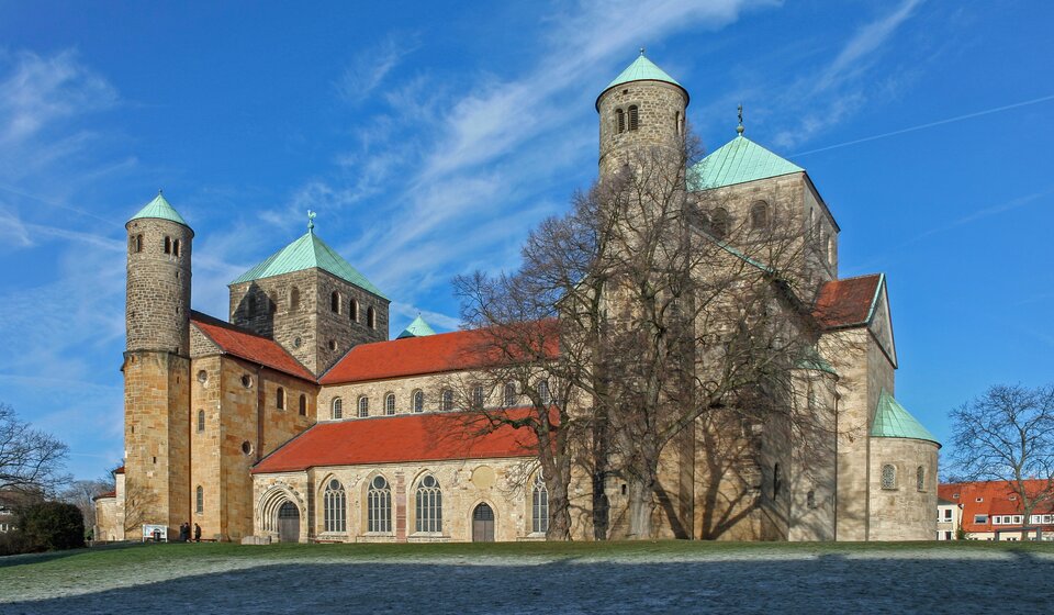 Zdjęcie przedstawia Kościół św. Michała w Hildesheim. Zdjęcie ukazuje budynek składający się z kilku brył o różnej wysokości, z dwoma transeptami i wieżami w typie ołówkowym. Na wszystkich ścianach świątyni znajdują się niewielkie otwory okienne zakończone półokrągłym łukiem. Na skrzyżowaniu naw znajduje się wieża na planie kwadratu. Dach korpusu budowli jest dwuspadowy.