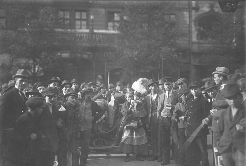 Zdjęcie przedstawia tłum ludzi na miejskiej ulicy. Wśród nich są głównie mężczyźni i młodzieńcy w kapeluszach i kaszkietach, a także jedna kobieta z małym dzieckiem na rękach. Pomiędzy ludźmi stoi pojazd. W tle widoczna jest kamienica oraz rosnące drzewa.