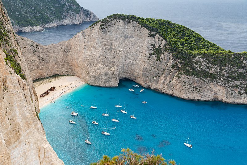 Zdjęcie z lotu ptaka przedstawia malowniczą plażę położoną w zatoczce, którą otaczają skały porośnięte u szczytu lasem. Woda ma kolor lazurowy. W pobliżu brzegu przycumowane są białe żaglówki ze spuszczonymi żaglami. Na plaży pokrytej jasnokremowym piaskiem widać drobniutkie ciemne punkciki – najprawdopodobniej to ludzie, oraz duży brązowy statek przypominający wrak. Na trzecim planie, za skałami widoczne jest szaroniebieskie morze, a po lewej stronie kadru – fragment kolejnej skały.