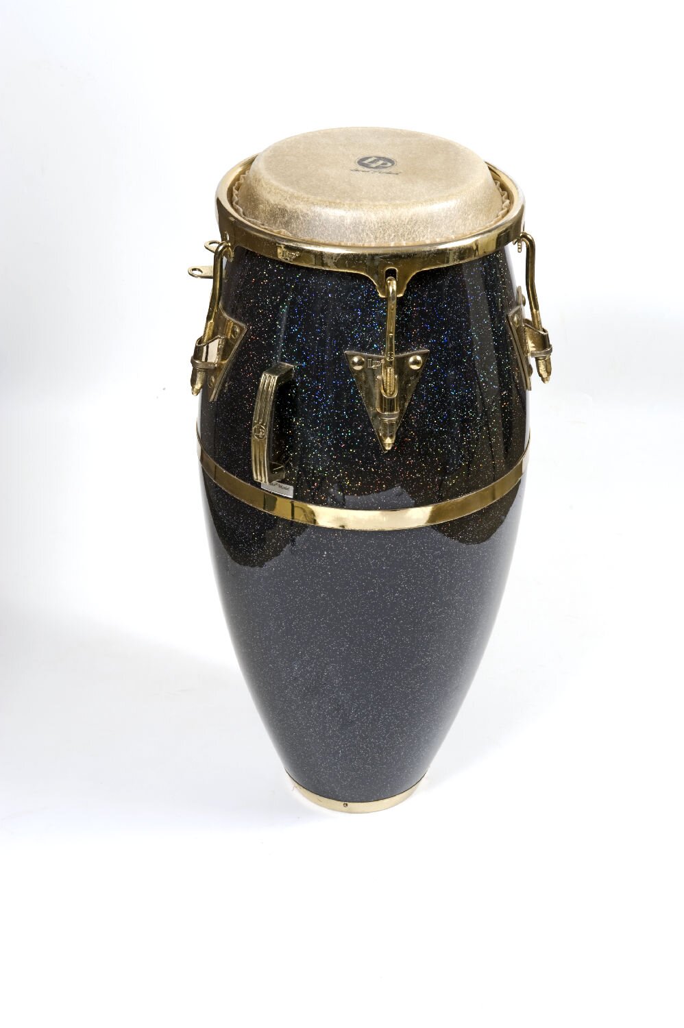 Zdjęcie przedstawia kongi w ujęciu z boku, pod lekkim kątem. Widoczny jest beczułkowaty kształt instrument zbudowany z czarnego beczułkowatego korpusu, na którym znajduje się szaro‑biała okrągła membrana wykonana ze skóry. Wokół membrany biegnie metalowa złota obejma, od której odchodzą pionowe, metalowe, złote elementy dotykające metalowej złotej obejmy biegnącej wokół korpus w centralnej części.