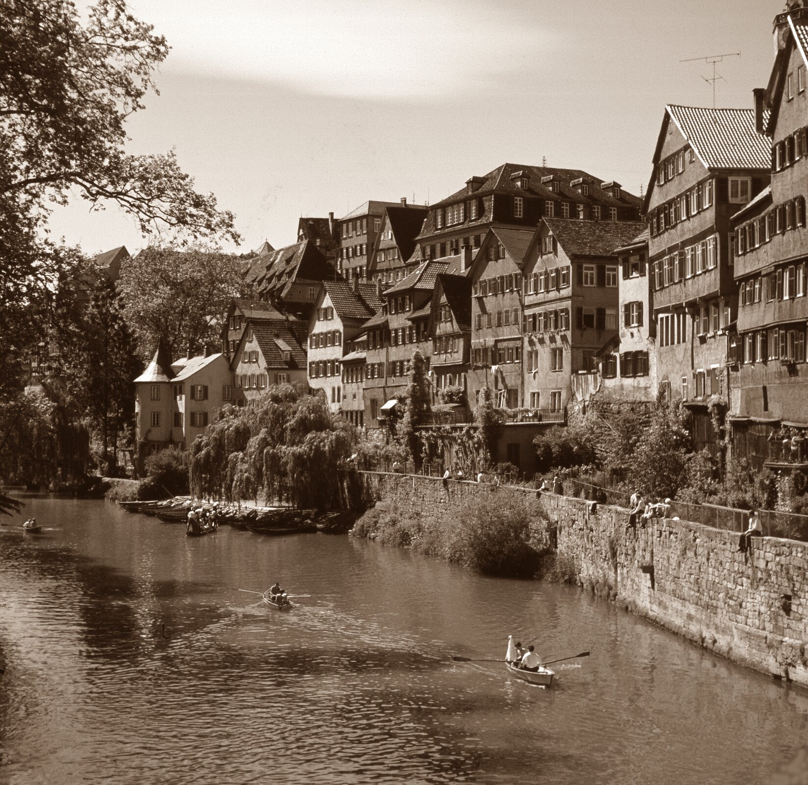 Ilustracja przedstawia fotografię wykonaną w sepii. Ukazuje widok na rzekę, płynącą przez Tybingę w Niemczech. Na brzegu znajdują się ciasno, jeden przy drugim dwu i trzypiętrowe budynki. Rzekę od budynków oddziela ceglany i kamienny mur, na jego szczycie siedzą przechodnie, patrząc na rzekę. Po wodzie łódkami płyną ludzie. Przy brzegu zacumowane są inne łodzie. Przy murze rosną krzaki i wierzby płaczące.