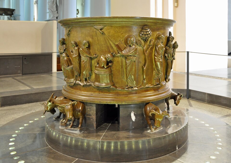 Romańska chrzcielnica z brązu: cylindryczna misa ozdobiona reliefami z postaciami biblijnymi, wsparta na figurach wołów; obiekt stoi w jasnym wnętrzu kościoła lub muzeum, na kamiennej posadzce.