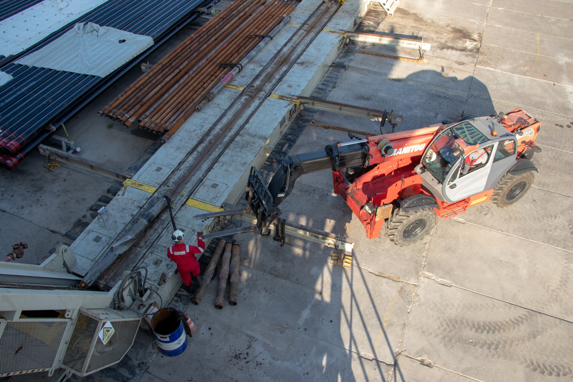 Na zdjęciu widoczny jest pomost rurowy i ześlizg. Po lewej stronie zdjęcia widać metalową pochylnię. Pochylnia zakończona jest na dole pomostem rurowym. Pomost jest w kształcie długiego prostokąta środkiem którego biegnie zagłębienie. W zagłębienie wkładana jest rura. Po lewej stronie pomosty leżą długie rury. Po prawej stronie pomostu znajduje się pojazd posiadający specjalny wysięgnik przy pomocy którego może podnieść rurę. Przed pojazdem znajduje się metalowy stelaż. Rama stelaża jest o kształcie graniastosłupa o podstawie trójkąta.Przy połączeniu ześlizgu z pomostem stoi pracownik ubrany w czerwony strój roboczy i biały kask.