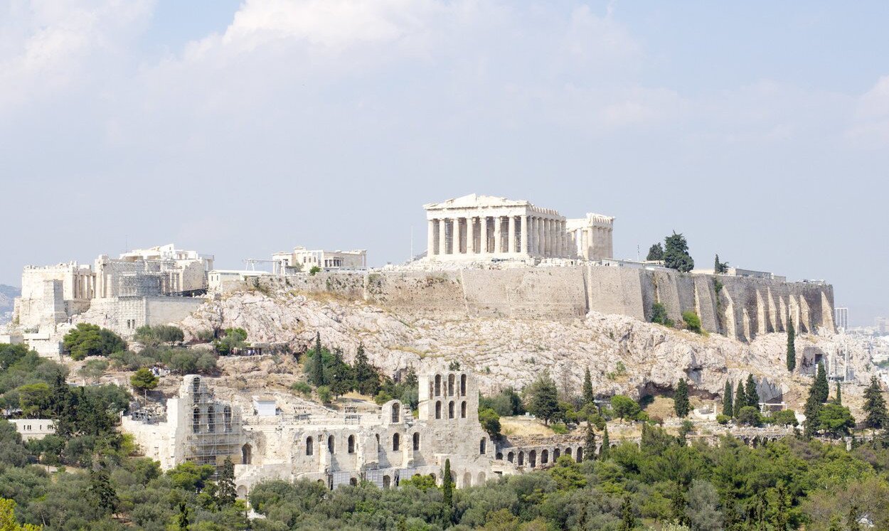 Fotografia prezentuje ruiny świątyń i pozostałości innych budowli na wapiennym wzgórzu Akropolu. Najbardziej widoczna ruina stojąca na szczycie wzgórza ma kształt prostopadłościanu. Konstrukcję ruiny podtrzymują stojące w rzędzie kolumny. Wzgórze obleczone jest grubym murem. Wzdłuż muru i skał widoczne są inne ruiny innych budowli, pomiędzy którymi rosną drzewa