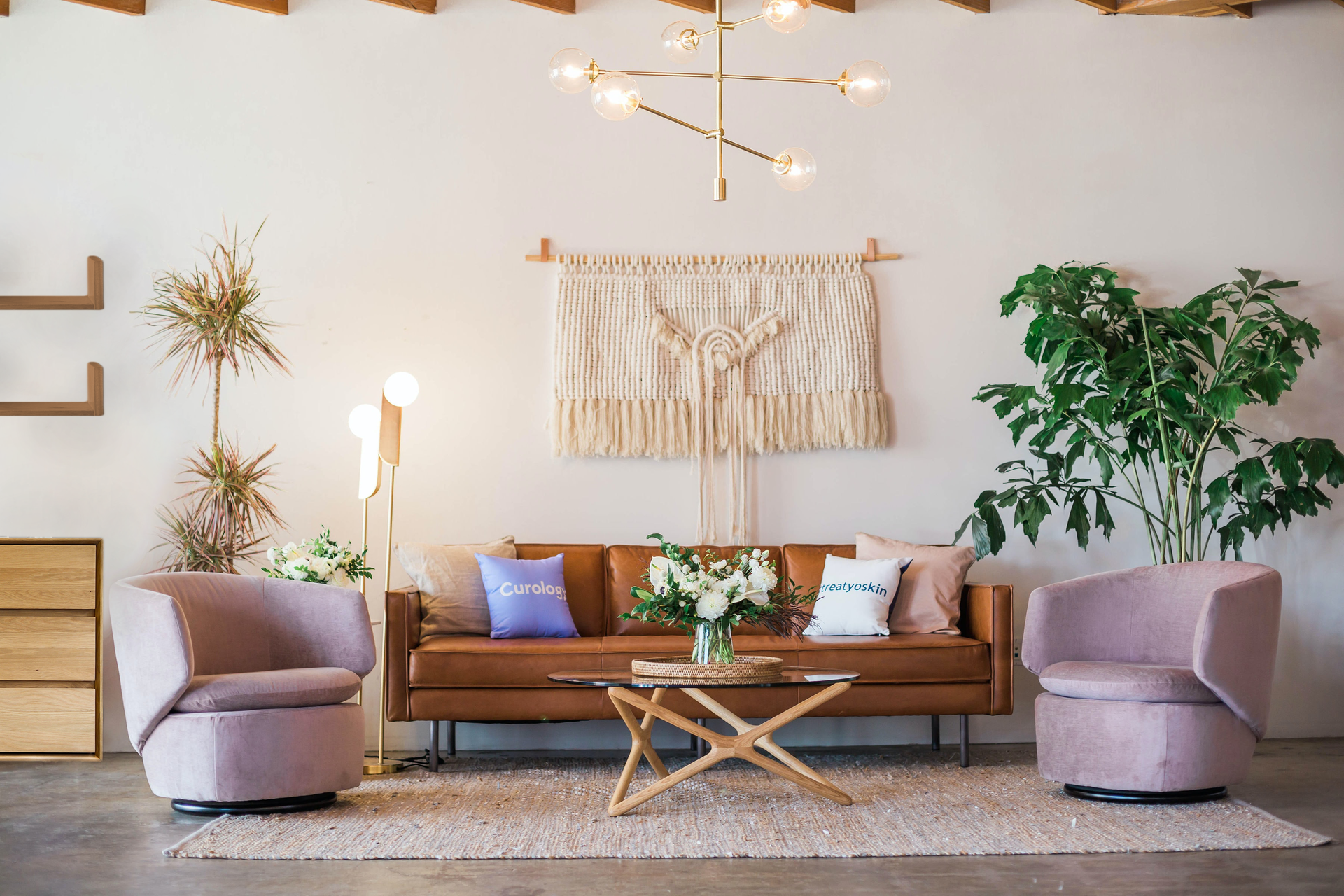 Zdjęcie przedstawia pokój. W centralnym miejscu stoi sofa. Przed sofą leży dywan, na którym stoi stolik i dwa fotele. Na stoliku stoją kwiaty w wazonie. Po prawej stronie sofy stoi kwiat w doniczce. Po lewej stronie sofy stoi lampa, kwiaty w doniczkach oraz komoda. Nad komodą wiszą dwie półki. Nad sofą, na ścianie wisi pleciona z włóczki makrama. Z sufitu zwisa żyrandol. 