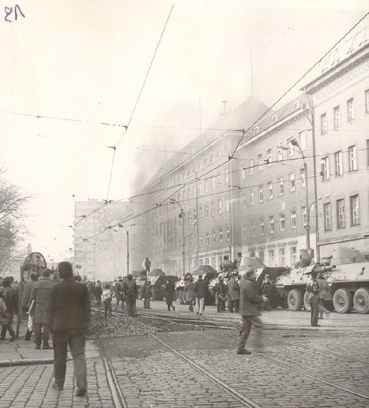 Zdjęcie przedstawia plac, na którym gromadzą się protestujący ludzie. Na placu stoi kolumna opancerzonych transporterów z żołnierzami.