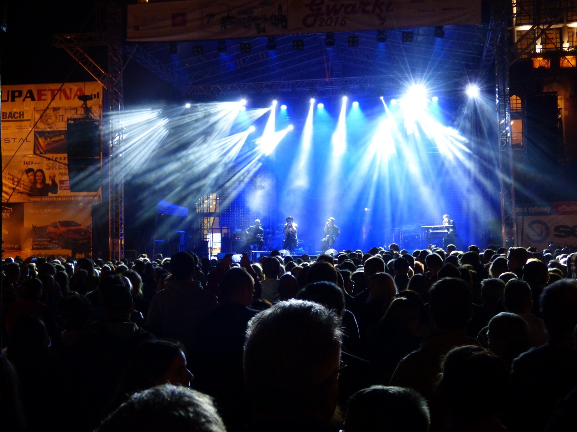 Zdjęcie przedstawia koncert muzyczny. Na pierwszym planie widać tłum ludzi stojących przed sceną, na drugim planie scenę rozświetloną reflektorami i niebieskim światłem. Na scenie znajduje się czterech muzyków. Za sceną widać fragmenty budynków.