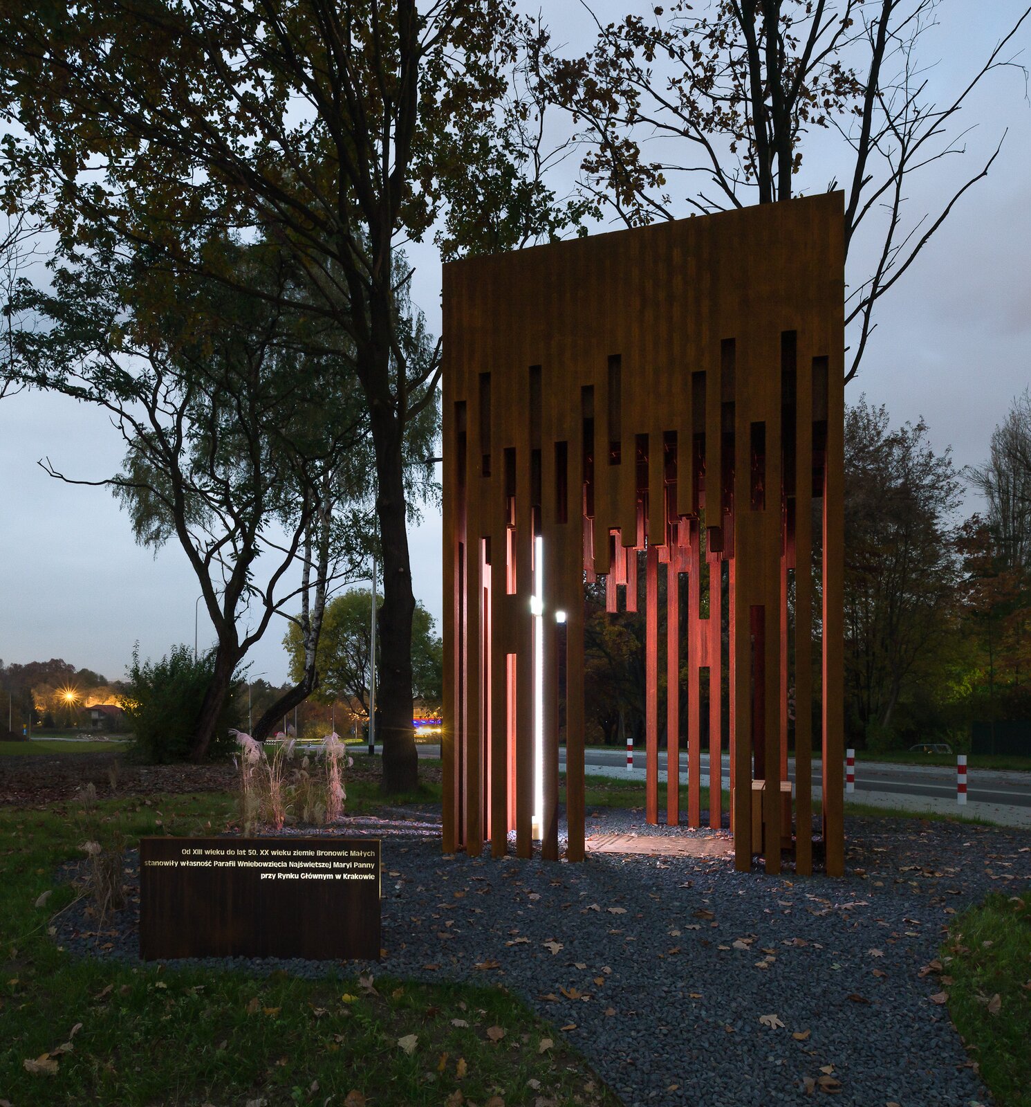 Fotografia przedstawia kapliczkę w Bronowicach Małych w bardzo nowoczesnym stylu. Konstrukcja jest wykonana z ciemnego drewna i ma prostopadłościenny kształt. Na górze jest w pełni zabudowana, a im niżej, tym więcej jest przestrzeni pomiędzy drewnianymi elementami. Elementy jak i przestrzenie między nimi mają kształt długich paneli. Z dwóch stron kapliczki znajdują się wysokie wejścia. W środku stoi wysoki świecący krzyż. Naprzeciwko niego znajduje się mała drewniana ławka. Przed wejściem do kapliczki, na kamiennym podłożu stoi tabliczka z podświetlanym napisem: "Od XIII wieku do lat 50. XX wieku ziemie Bronowic Małych stanowiły własność Parafii Wniebowzięcia Najświętszej Maryi Panny przy Rynku Głównym w Krakowie". Za kapliczką znajdują się drzewa i inna roślinność. Zdjęcie zrobiono o zmroku.