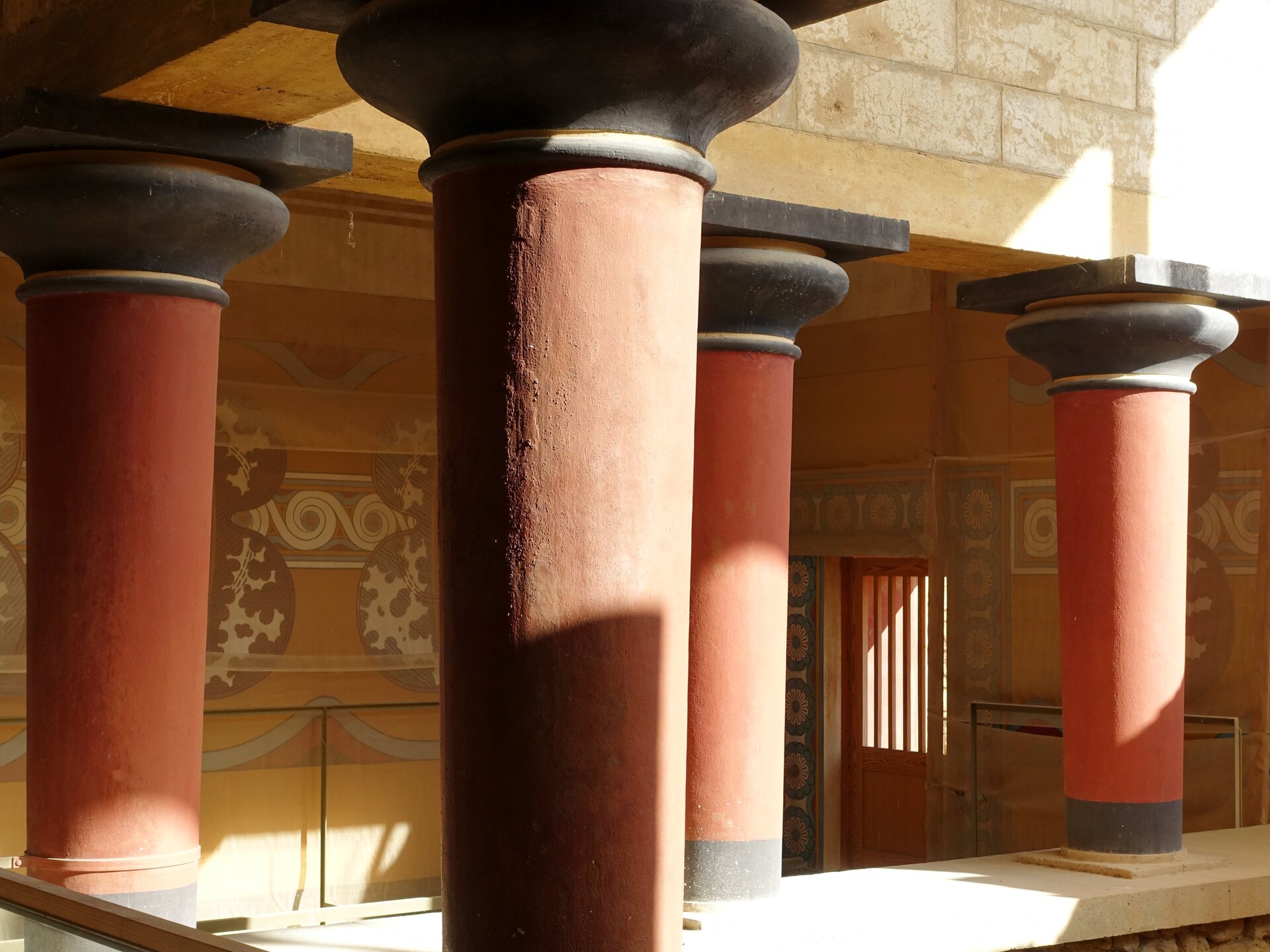 Ilustracja przedstawia salę kolumnową w pałacu w Knossos. Na fotografii ukazano cztery czerwone kolumny o czarnych głowicach. Wspierają one strop. Na żółtym tle ściany namalowany jest pas dekoracji.