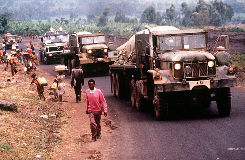 Zdjęcie przedstawia kilka wojskowych ciężarówek, które jadą po drodze. Po lewej stronie, na poboczu drogi, idą czarnoskórzy mężczyźni i kobiety. Część niesie ze sobą baniaki. W tle widoczne są drzewa.
