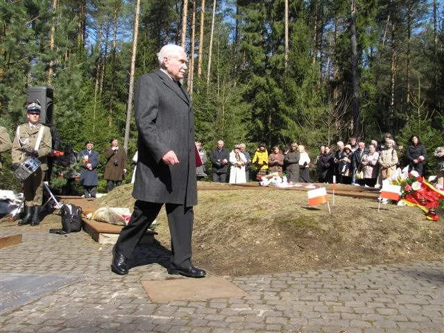 Kliknij, aby powiększyć Zdjęcie przedstawia idącego starszego mężczyznę, z siwymi włosami i wąsami. Jest ubrany w jesienny płaszcz. Za nim znajduje się niewielki kopiec, na którym leżą kwiaty i wbite są chorągiewki z polską flagą. Za kopcem stoją ludzie. Na lewo od mężczyzny widać polskiego żołnierza w mundurze.