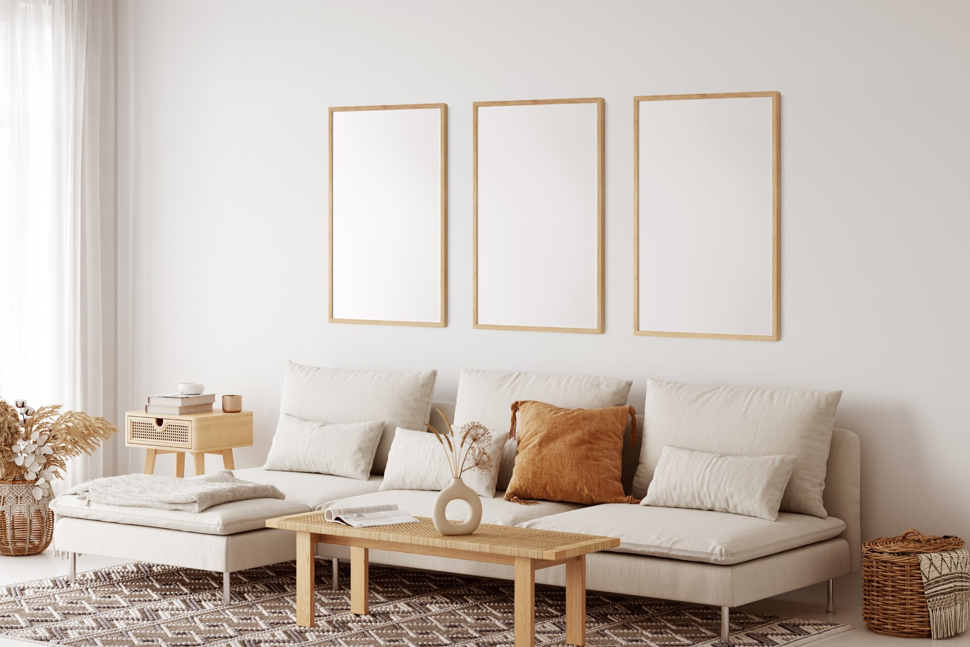 Fotografia przedstawia widok na pomieszczenie urządzone w stylu skandynawskim, które cechuje się jasnymi ścianami i niezwykłą prostotą. Pośrodku stoi jasna sofa, na której ułożono różnej wielkości poduszki – jedna z nich jest pomarańczowa, a pozostałe beżowe. Przed meblem widać niski stolik w odcieniu ciepłego drewna, na którym stoi dekoracyjna waza z pustym okręgiem w środku. Z lewej strony fotografii widać drewnianą szafkę z małą szufladą, na której ułożono książki oraz pojemnik na świecę oraz pojemnik na liście dekoracyjne stojący na jasnej podłodze. Za nim widać długie, białe zasłony. Z Prawej strony sofy widać kosz wiklinowy z przykrywką, z którego wystaje kawałek materiału przypominający koc. W środku pomieszczenia leży dywan w geometryczne wzory w odcieniach szarości, brązu i bieli. Na ścianie za meblem widać trzy obrazy w drewnianej oprawie, które przedstawiają jedynie białą powierzchnię. 