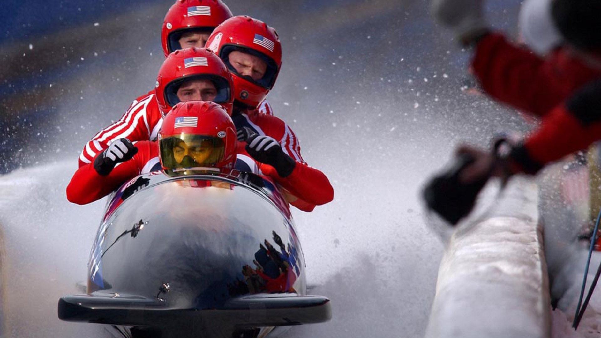 Zdjęcie przedstawia lodowy tor w kształcie rynny. Na torze znajdują się  sanie bobslejowe z czterema mężczyznami w sportowych strojach i kaskach. Za bobslejem rozpyla się mgła kruszonego lodu.
