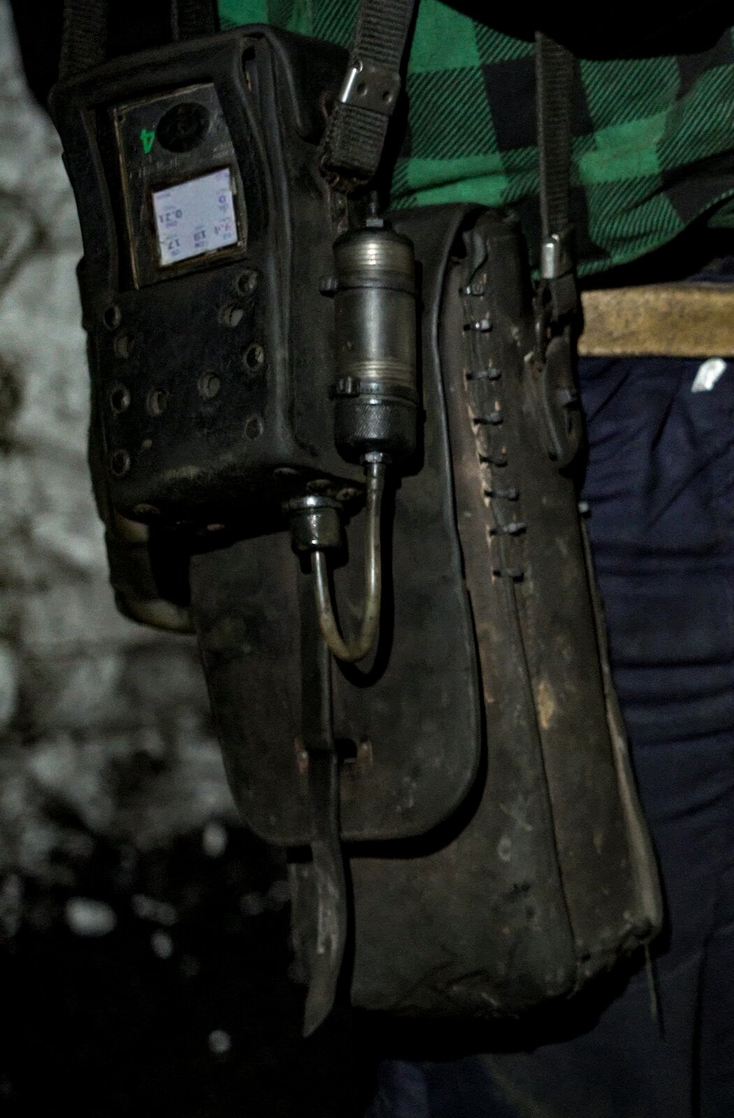 Zdjęcie przedstawia detektor wielogazowy, będący na wyposażeniu górnika. Detektor jest urządzeniem o kształcie prostopadłościanu. Na przedniej ścianie prostopadłościanu widoczny jest ekran. Od dolnej podstawy odchodzi rurka. Rurka łączy się z małym elementem o kształcie cylindra. Cylinder jest zawieszony na bocznej ścianie detektora. Detektor znajduje się w pokrowcu. Pokrowiec jest zawieszony na pasku spodni górnika. 