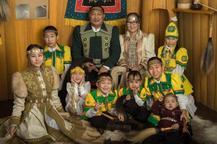 Rodzina Jakutów. Na zdjęciu dziesięcioosobowa rodzina. Kobieta , mężczyzna, trzy dziewczynki i sześciu chłopców. Część z nich ma na sobie żółto zielone stroje. Kobieta i najstarsza córka mają na sobie brązowe stroje. Mężczyzna ciemno‑zielony strój. 