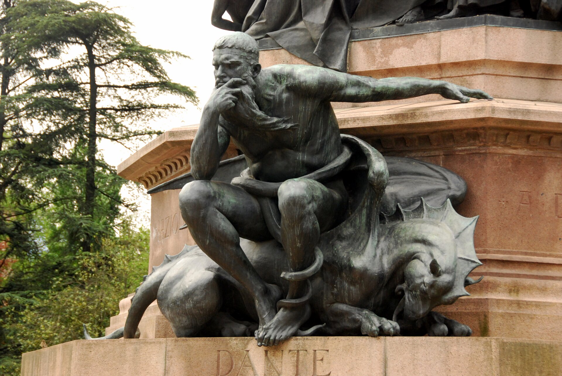 Zdjęcie przedstawia ciemnoszarą rzeźbę mężczyzny. Jest on nagi, siedzi na smoku, którego ogon zawija się wokół jego tułowia. Mężczyzna prawą dłonią przykłada do długiej brody, a lewą wysunął za siebie, podpierając się lekko.