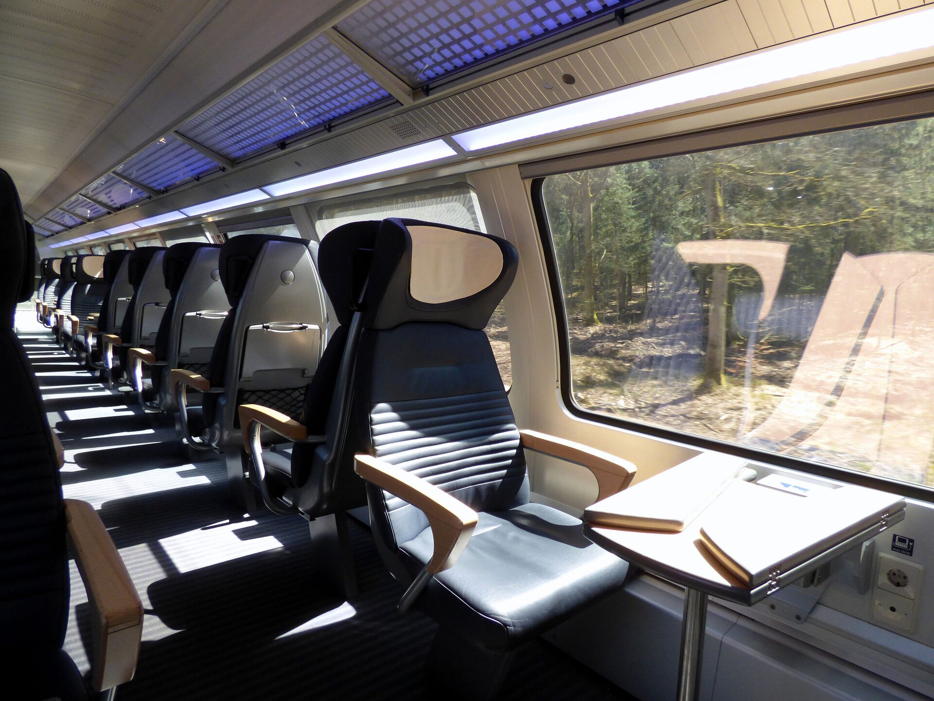 Zdjęcie przedstawia wnętrze wagonu pociągu z rzędem foteli ustawionych jeden za drugim. Nad siedzeniami umieszczona jest półka bagażowa. Widoczne są szerokie okna wagonu.