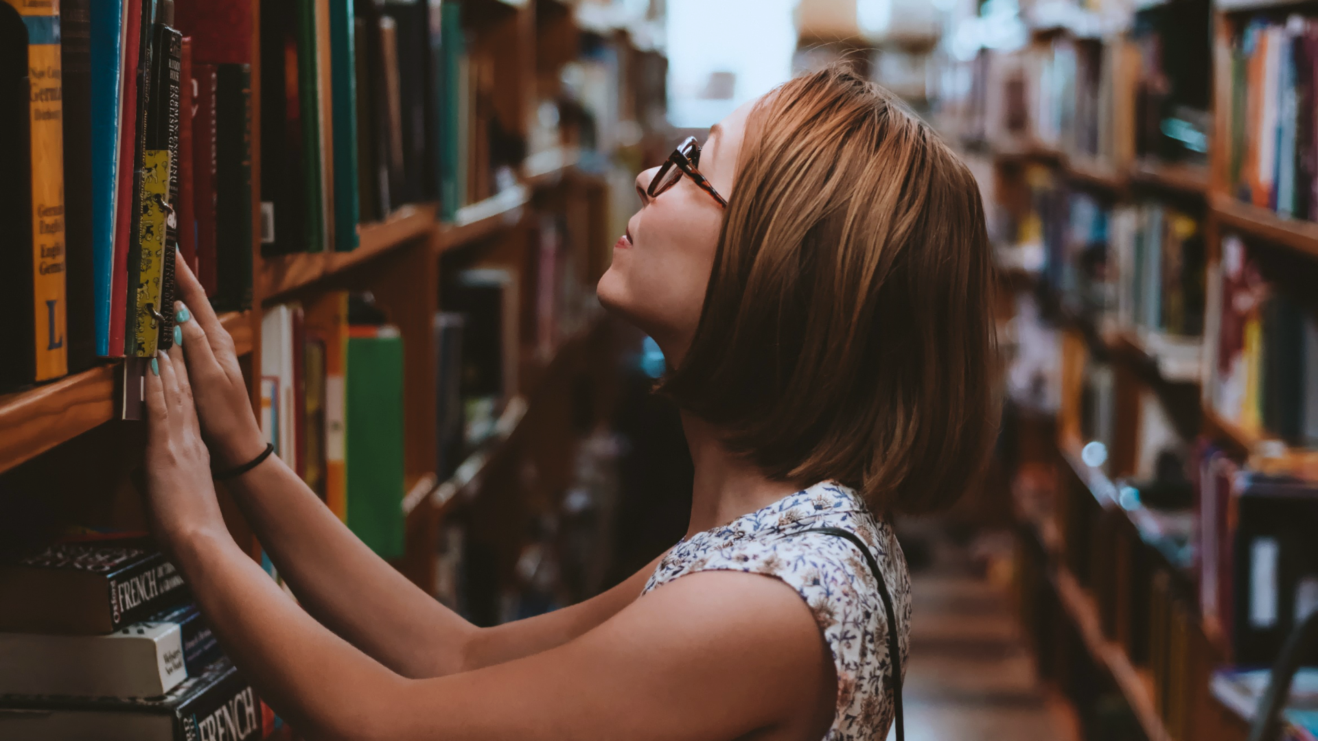 Zdjęcie przedstawia kobietę w okularach przeglądającą książki na półce w bibliotece.