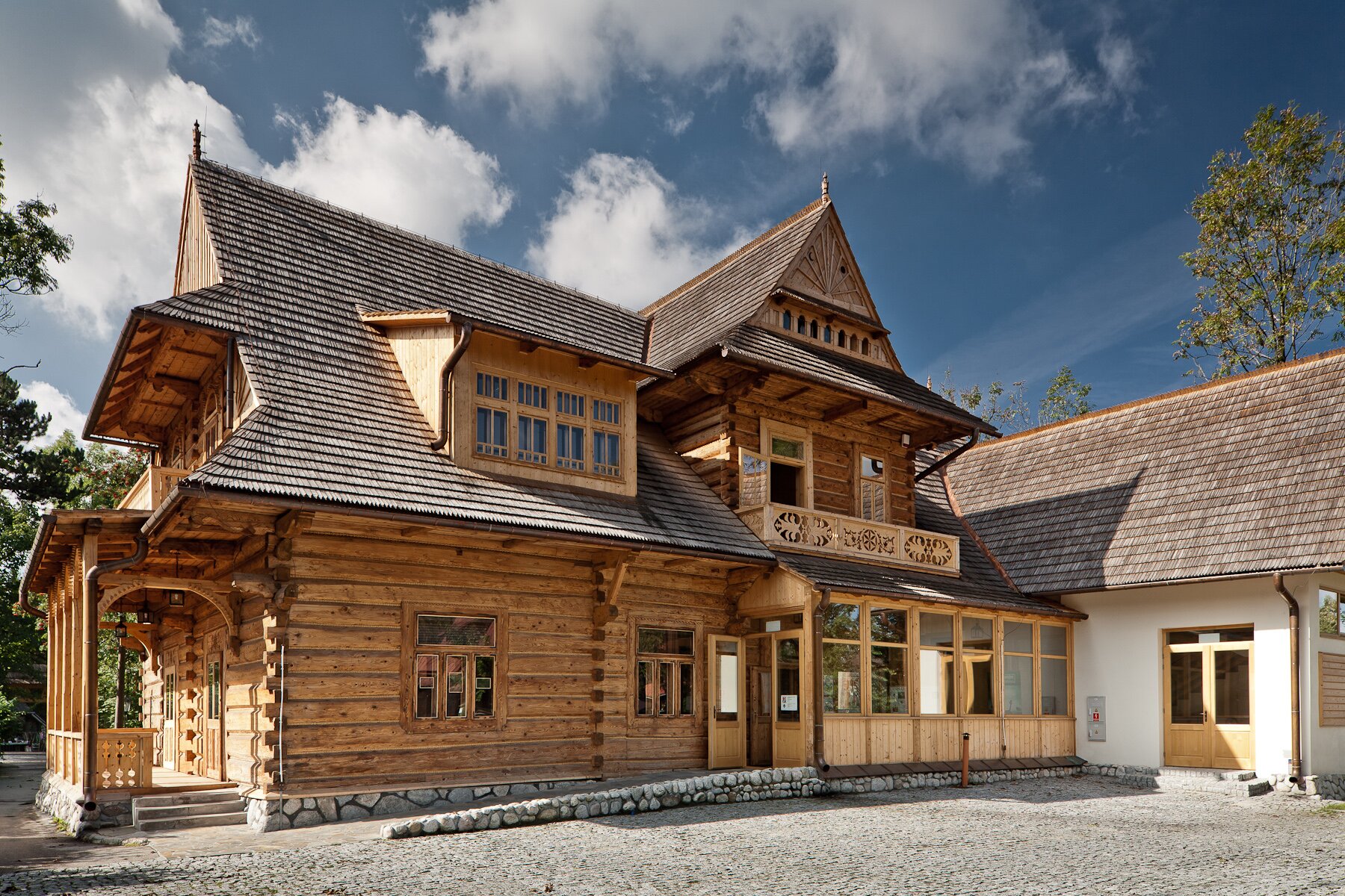 Fotografia okazałego, drewnianego budynku. Kolor ścian jasnobrązowy, spadzisty dach w kolorze ciemnoszarym. Weranda, u góry balkony.