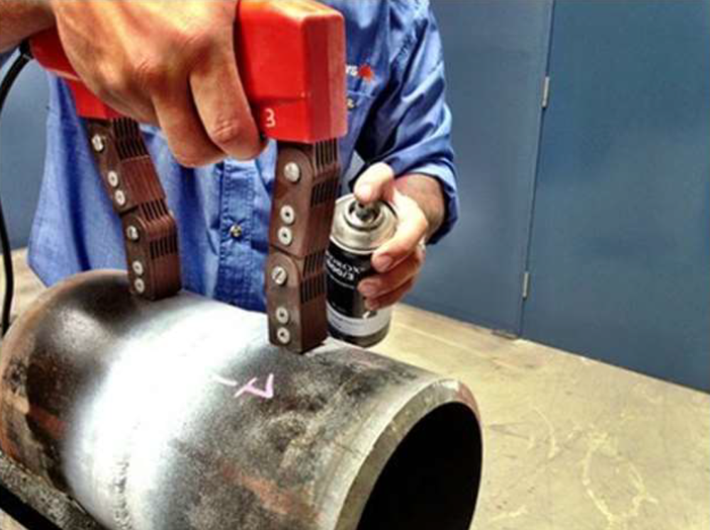 Na zdjęciu znajduje się mężczyzna, który trzyma w dłoni magnes przyłożony do obiektu, a w drugiej dłoni puszkę z proszkiem fluorescencyjnym, za pomocą których prowadzi badanie w celu wykrycia pęknięć i defektów.