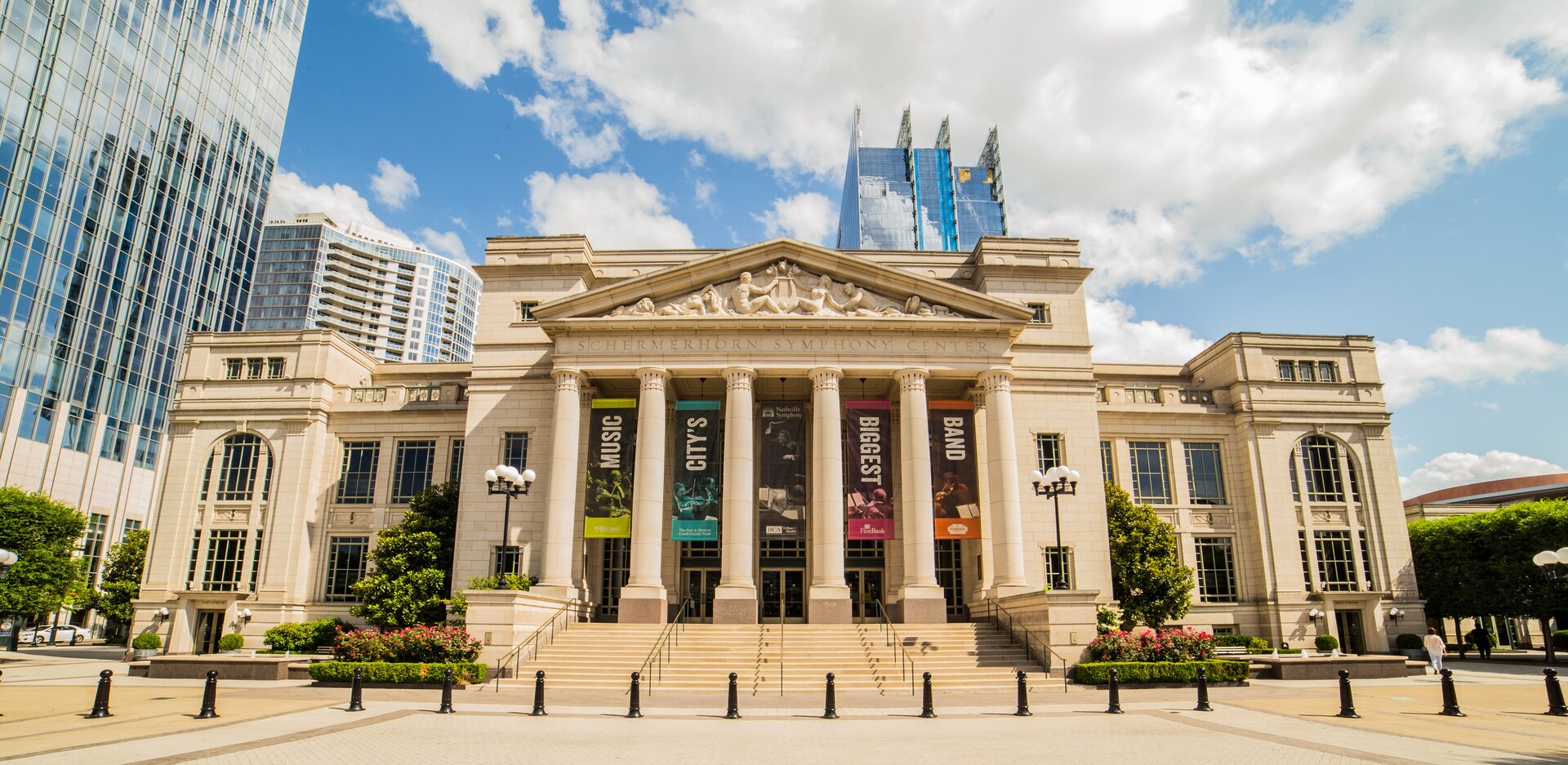 Ilustracja przedstawia Schermerhorn Symphony Center w Nashville w USA. Budynek widoczny jest od frontu. Przed wejściem znajdują się wysokie kolumny. W tle widoczne są nowoczesne, oszklone budynki.