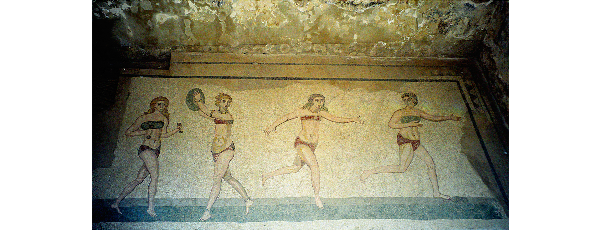 Zdjęcie przedstawia mozaiki na ścianie w Villa Romana del Casale. Mozaika przedstawia cztery kobiety wykonujące ćwiczenia sportowe.