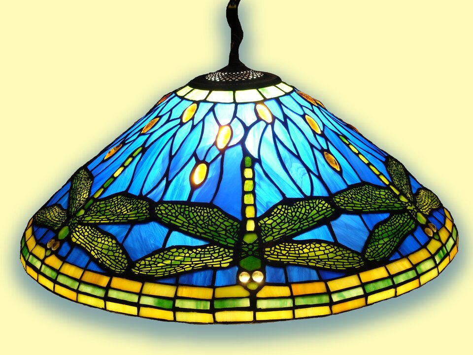 Ilustracja przedstawia fotografię lampy. Lampa ma kształt stożka o szerokiej podstawie i ściętego u nasady, w której zamocowany jest kabel do zawieszenia lampy na suficie. Lampa jest wykonana z kawałków kolorowych szkieł w różnych odcieniach złotego, zielonego i niebieskiego. . Lampa jest ozdobiona zielonymi wizerunkami ważek, które lecą po niebieskim niebie. Ważki są ukazane na wprost, nieco z góry i stanowią powtarzalny element umieszczony na dole, wokół całej lampy..