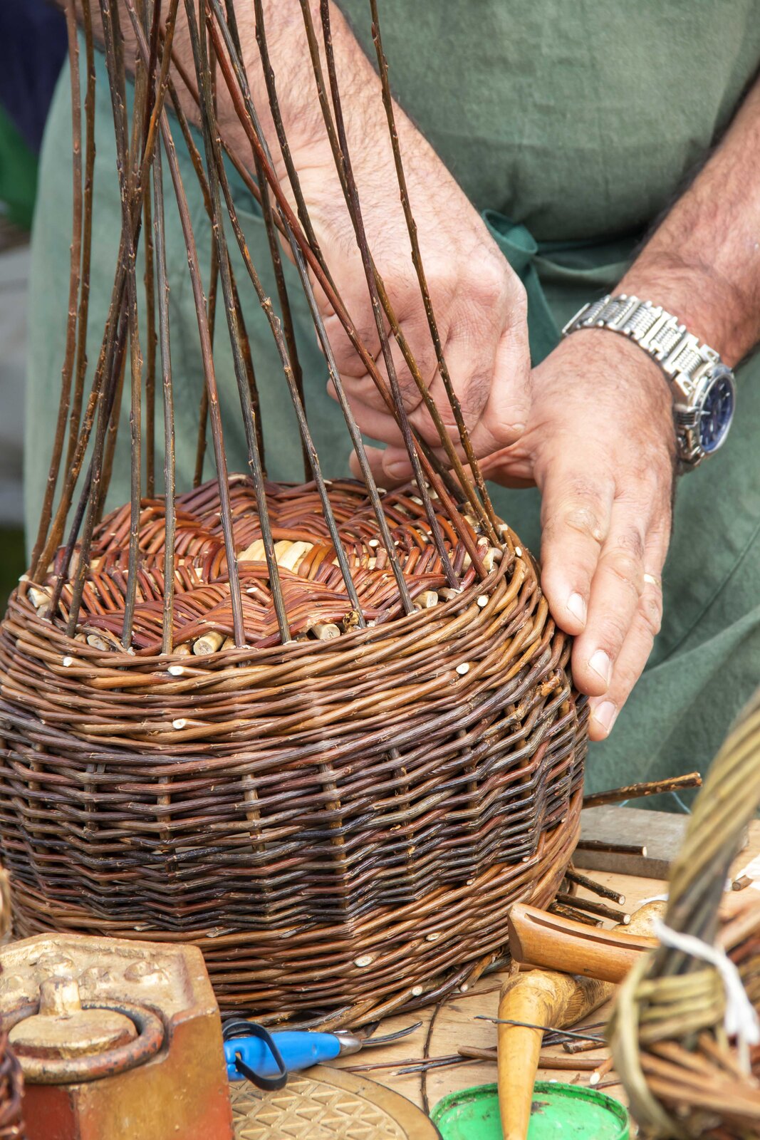 Zdjęcie kosza zielonego, czyli wyrobu plecionkarskiego z wikliny nieokorowanej. Prezentowany wyrób ma okrągłe dno i jest w kolorze ciemno i jasnobrązowym. Na zdjęciu widoczne są dłonie koszykarza‑plecionkarza, który wyplata wykończenie kosza.
