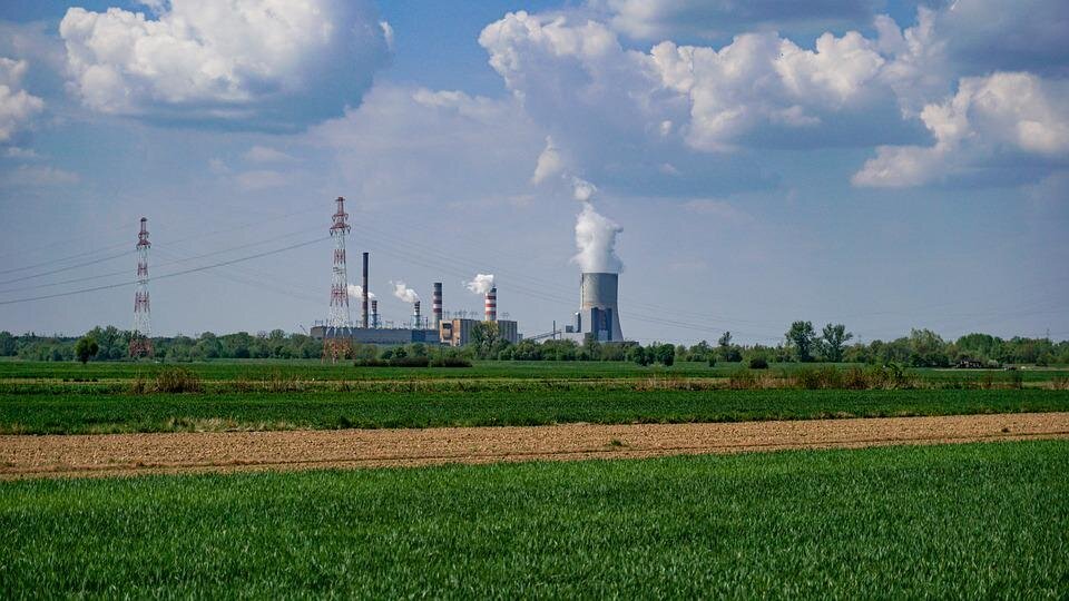 Na zdjęciu Elektrownia Kozienice. W oddali kilka dużych budynków, wokół których zbudowano ogromne kominy. Wokół pola uprawne.