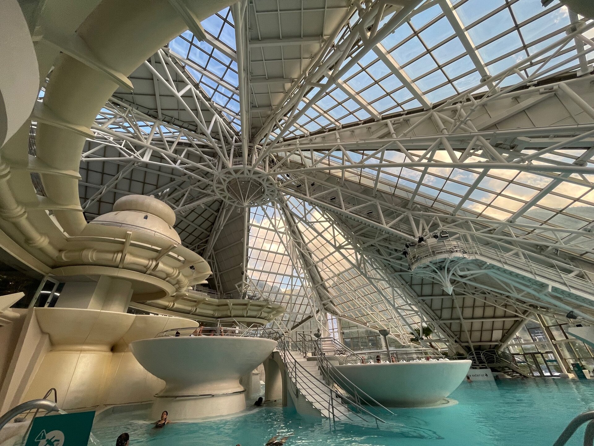 Fotografia przedstawia fragment wnętrza obiektu rekreacyjnego: kompleksu basenów krytych szklaną kopułą. Większość kadru zajmuje konstrukcja - metalowy szkielet, na który składa się wiele prostych elementów krzyżowanych ze sobą. Pomiędzy gęstą siecią elementów konstrukcyjnych znajdują się wielkie otwory okienne, doświetlające przestrzeń nad basenami.
