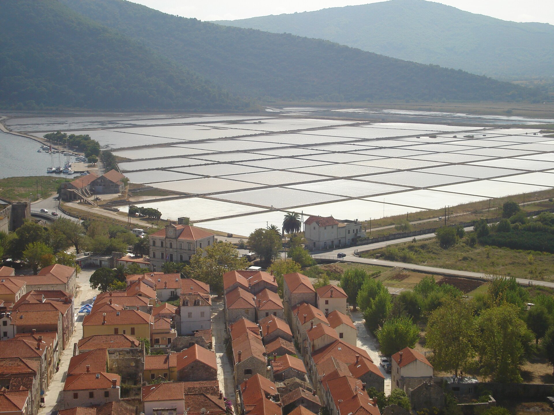 Na zdjęciu jest salina. To część terenu zalana wodą, podzielona na kwadratowe pola. Na pierwszym planie stoją domy. W tle są wzniesienia porośnięte drzewami.