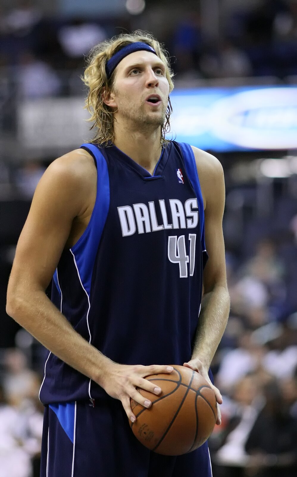 Zdjęcie przedstawia Dirka Nowitzkiego. To młody mężczyzna w sportowej koszulce, spodenkach i przepasce na włosach. Mężczyzna ma długie blond włosy. Jego twarz jest pociągła. Mężczyzna trzyma w rękach piłkę do koszykówki.