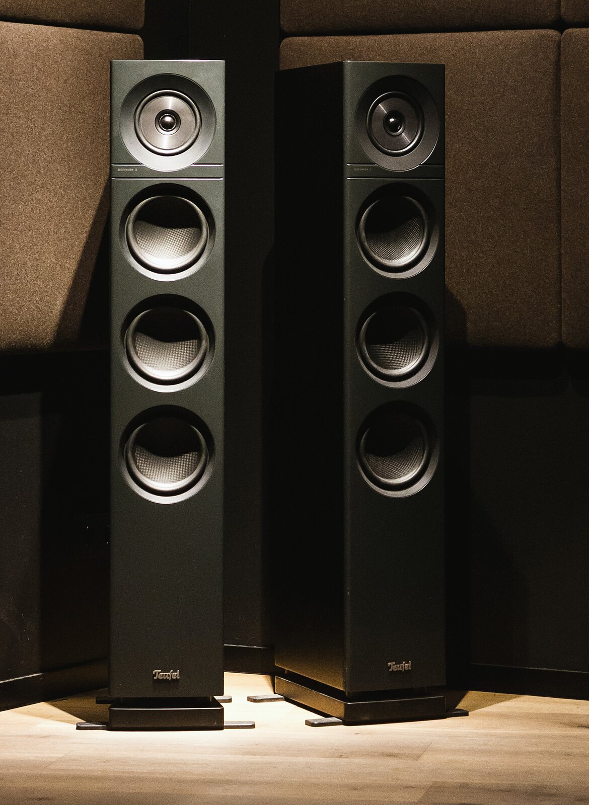 Na zdjęciu widoczne są dwa głośniki kolumnowe w pomieszczeniu. Po lewej frontalnie ustawiony srebrny głośnik, w którego górnej części znajduje się okrągły element z kratownicą, poniżej którego znajdują się trzy okrągłe membrany. Głośnik jest ustawiony na czarnej prostopadłościennej podstawie. Po prawej czarny głośnik w ujęciu trzy czwarte, w którego górnej części znajduje się okrągły element z kratownicą, poniżej którego znajdują się trzy okrągłe membrany. Głośnik jest ustawiony na czarnej prostopadłościennej podstawie. Głośniki stoją na drewnianej podłodze w kolorze beżowym, zaś za głośnikami znajdują się duże, prostokątne, szare panele wygłuszające.