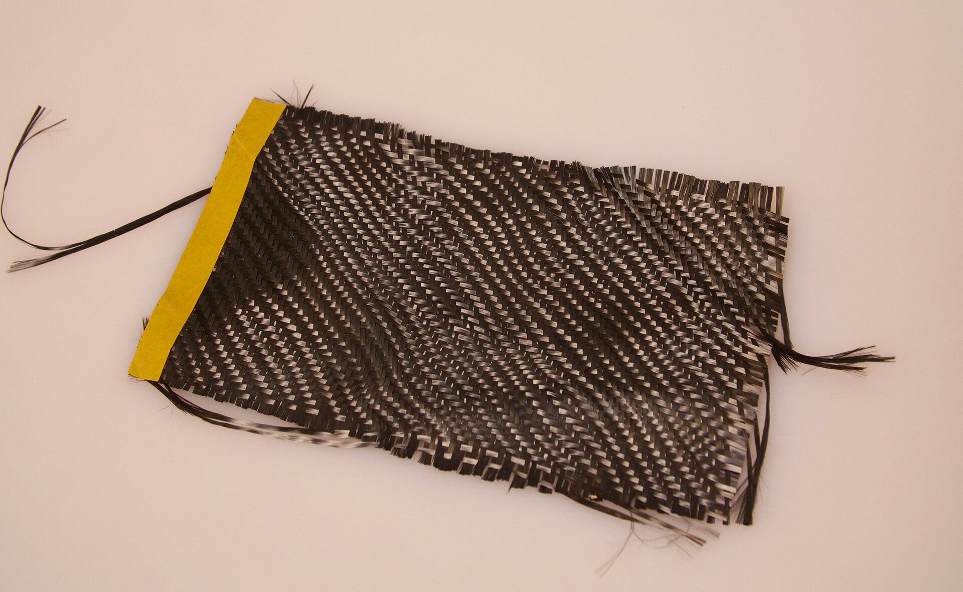 Zdjęcie przedstawia włókno węglowe. Wycinek materiału ma prostokątny kształt. Jest czarno‑szary, lekko połyskliwy. Posiada wzór w postaci ukośnie poprowadzonych pasów, naprzemiennie - jasnych i ciemnych. Z lewej strony posiada żółtą lamówkę. Z prawej brzeg materiału jest częściowo naddarty.