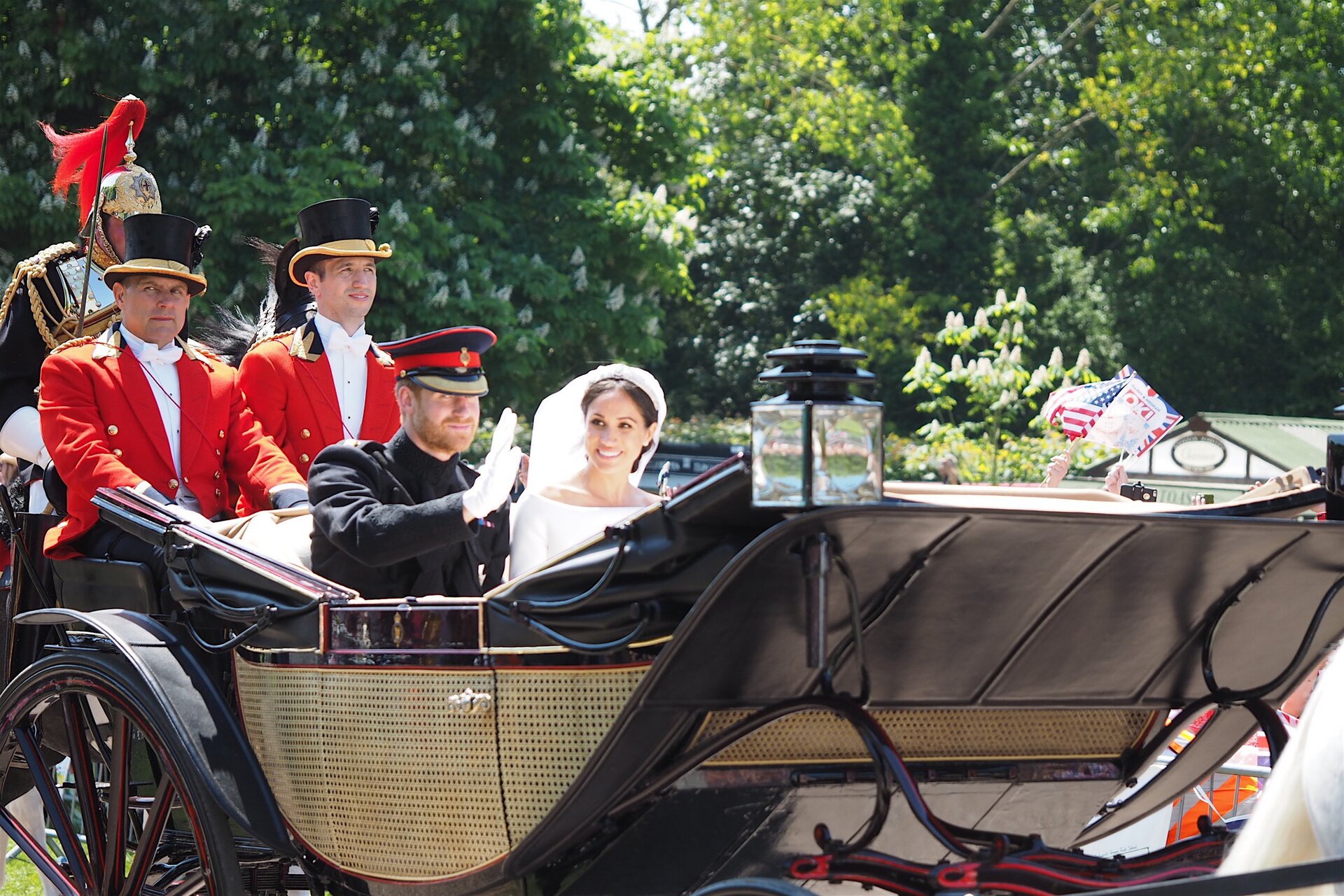 Fotografia barwna przedstawiająca księcia Harry'ego i Meghan Markle, jadących karocą. Książę - rudy, brodaty mężczyzna - ubrany jest w mundur i wojskową czapkę. Macha ręką w białej rękawiczce. Obok niego Meghan Markle, szczupła , długowłosa brunetka, w białej sukni z welonem na głowie. Oboje uśmiechnięci.