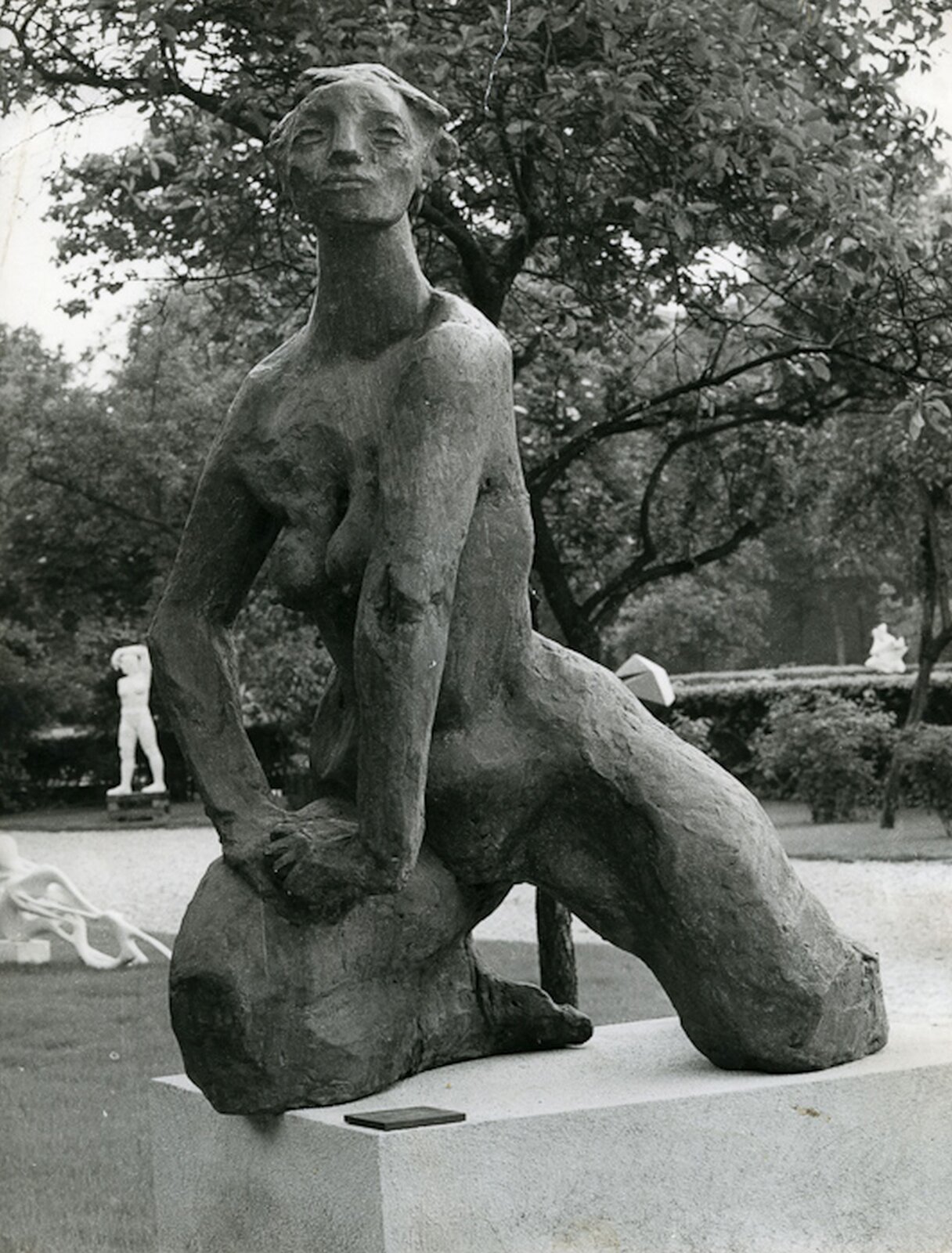 Zdjęcie przedstawia rzeźbę Aliny Szapocznikowej Pomnik dla spalonego miasta. Rzeźba przedstawia nagą kobietę. Kobieta klęczy na podłożu, dłonie obu rąk splotła na udzie prawej nogi. Rzeźba ustawiona jest w parku w tle widoczne są drzewa i krzewy.