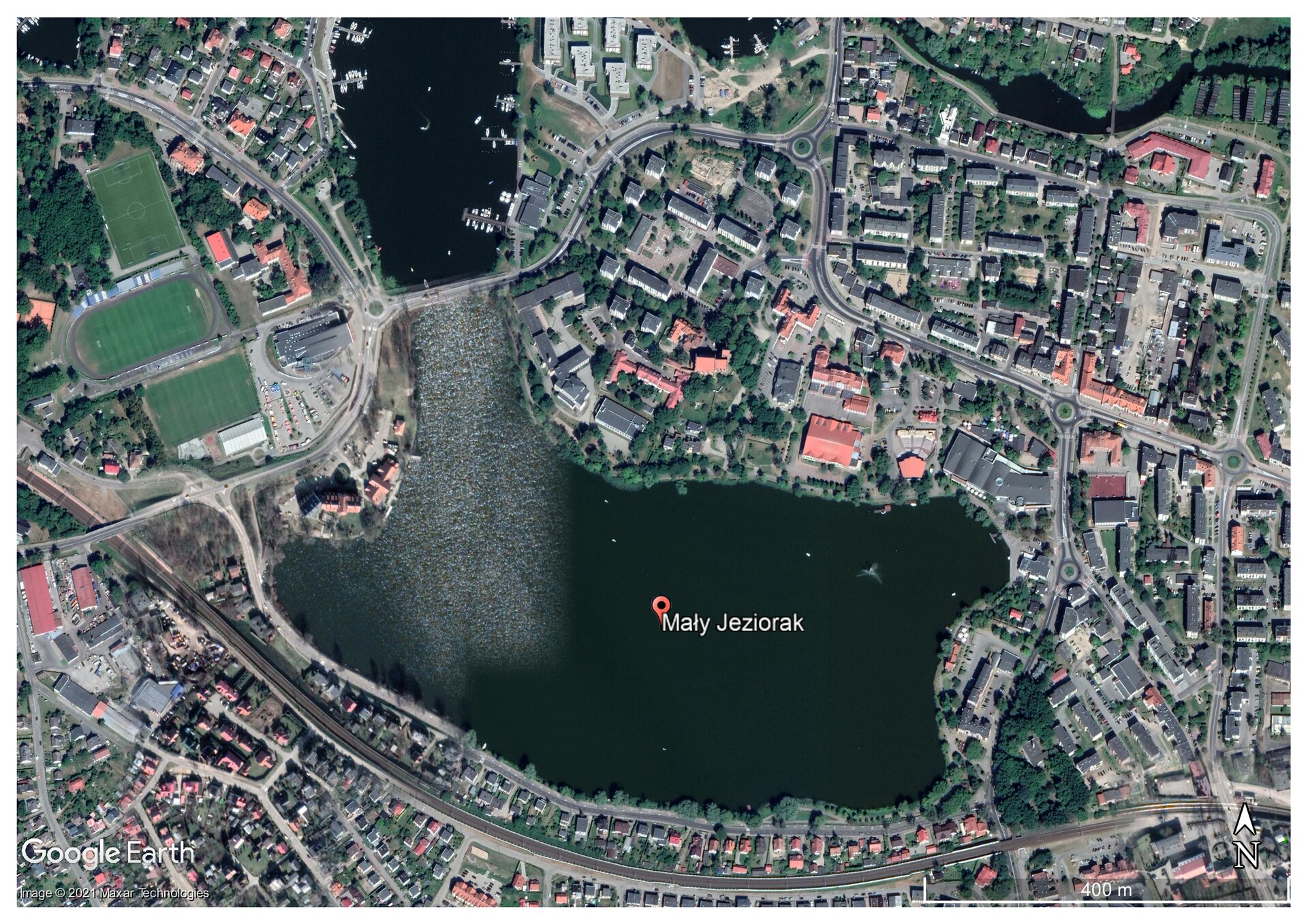 Zdjęcie satelitarne przedstawiające jezioro Mały Jeziorak. Znajduje się ono w centralnej części, pośrodku umieszczono czerwoną wirtualną pinezkę z nazwą jeziora. Woda jest bardzo ciemnego koloru, prawie czarnego. Po lewej stronie na tafli na biało odbijają się promieni słońca. Dookoła jeziora zabudowania miejskie. Gęsta zabudowa składająca się głównie z domów jednorodzinnych oraz większych budynków. W lewym górnym rogu trzy zielone boiska piłkarskie. Pomiędzy budynkami zieleń miejska, drogi. U dołu, równolegle do linii jeziora, biegnie linia kolejowa. Wysoki poziom szczegółów, można rozpoznać pojedyncze pojazdy stojące na parkingach. 