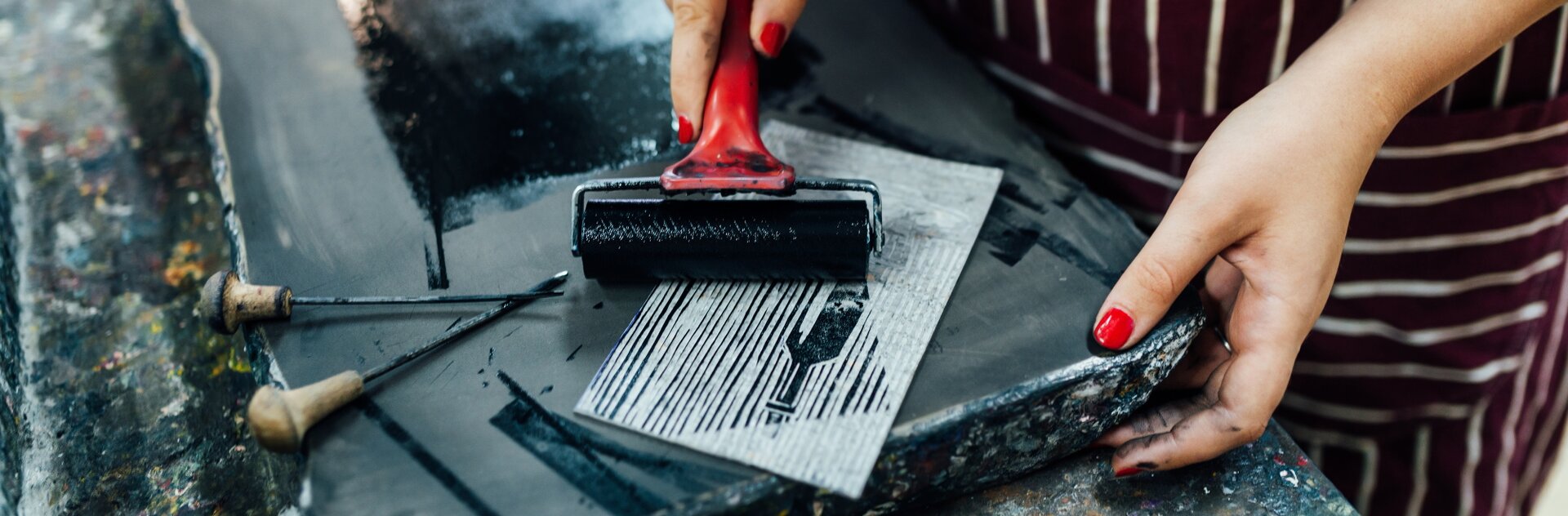 Fotografia przedstawia  stanowisko pracy oraz dłonie kobiety, która przy nim stoi. W prawej dłoni kobieta trzyma wałek o czerwonym trzonie, który został zanurzony w czarnej substancji. Kobieta, której paznokcie pomalowane są na czerwono, przeciąga wałek po wzorniku, zamalowując jego powierzchnię w czarne pasy. Ma na sobie fartuch w pasy. Po lewej stronie, na blacie przypominającym kamień leżą dwa narzędzia, różniące się stopniem zużycia, przypominające dłuta o cienkich zakończeniach i małych, drewnianych trzonach.
