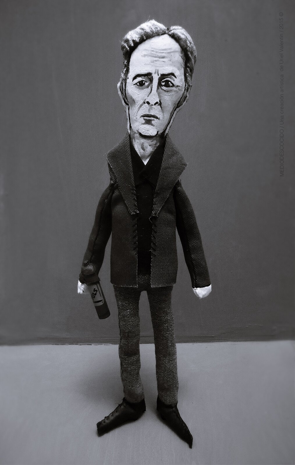 Zdjęcie przedstawia pacynkę w kształcie człowieka. Jest to dojrzały mężczyzna. Ma długą twarz ze zmarszczkami. Ma krótkie, siwe włosy. Ubrany jest w ciemny płaszcz. W prawej ręce trzyma butelkę.