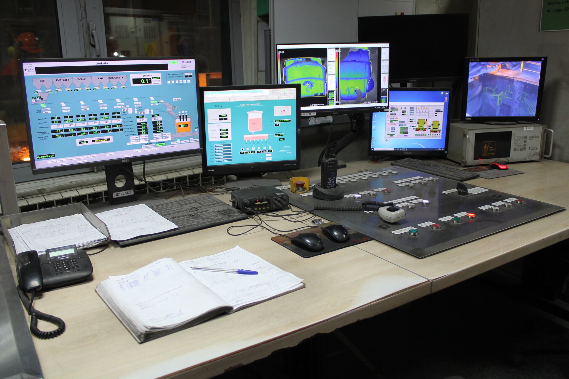 Zdjęcie przestawia Pulpit sterowniczy w kabinie operatorskiej na stanowisku ciągłego odlewania stali. Na zdjęciu widoczne jest biurko. Na biurku znajduje się sześć monitorów. Jeden z nich jest wyłączony, a pozostałe przedstawiają parametry stanowiska ciągłego odlewania stali. Na biurku znajduje się otworzony zeszyt z długopisem, dokumenty, telefon stacjonarny, klawiatura, dwie myszki na podkładce, krótkofalówka oraz pulpit sterowniczy. Pulpit składa się z kilkunastu przycisków w różnych kolorach. Każdy przycisk jest opisany. W tle widoczne jest okno, a za nim znajduje się stanowisko odlewania stali.