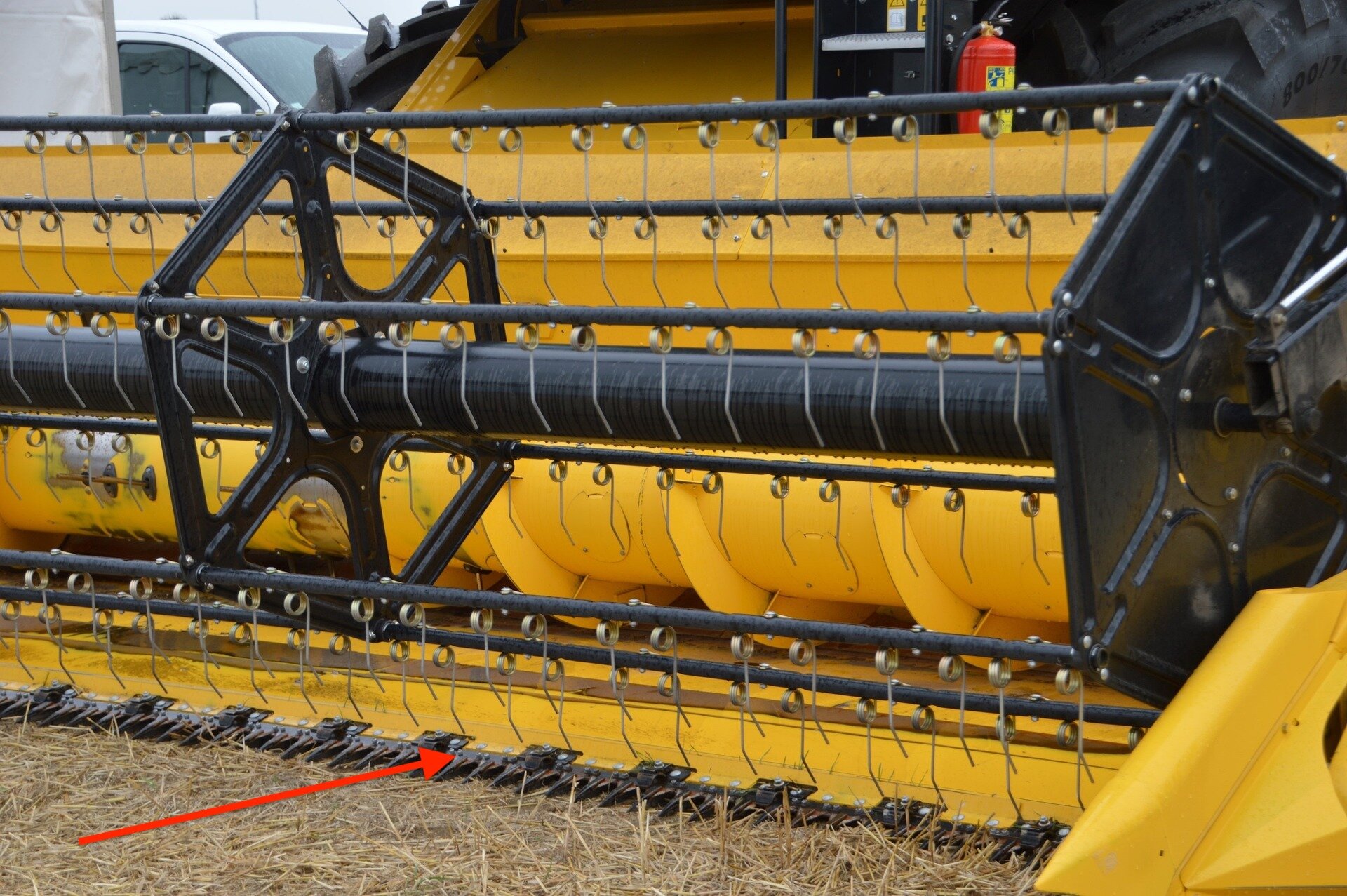 Zdjęcie przedstawia zbliżenie na heder kombajnu zbożowego. Czerwoną strzałką zaznaczono zespół tnący. Składa się on z nieruchomej belki z przymocowaną do niej ruchomą listwą nożową, wyposażoną w ostre nożyki. Do belki palcowej przymocowane są również palce zespołu tnącego. Mają podłużny kształt, zwężający się ku przodowi.