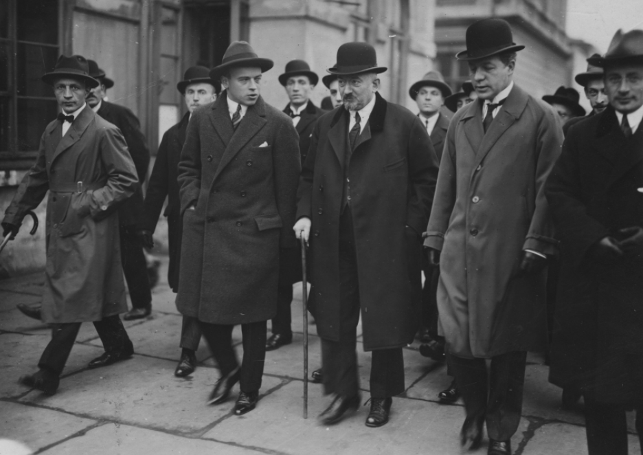 Na zdjęciu grupa mężczyzn w garniturach, płaszczach i kapeluszach. Grupa idzie po ulicy.