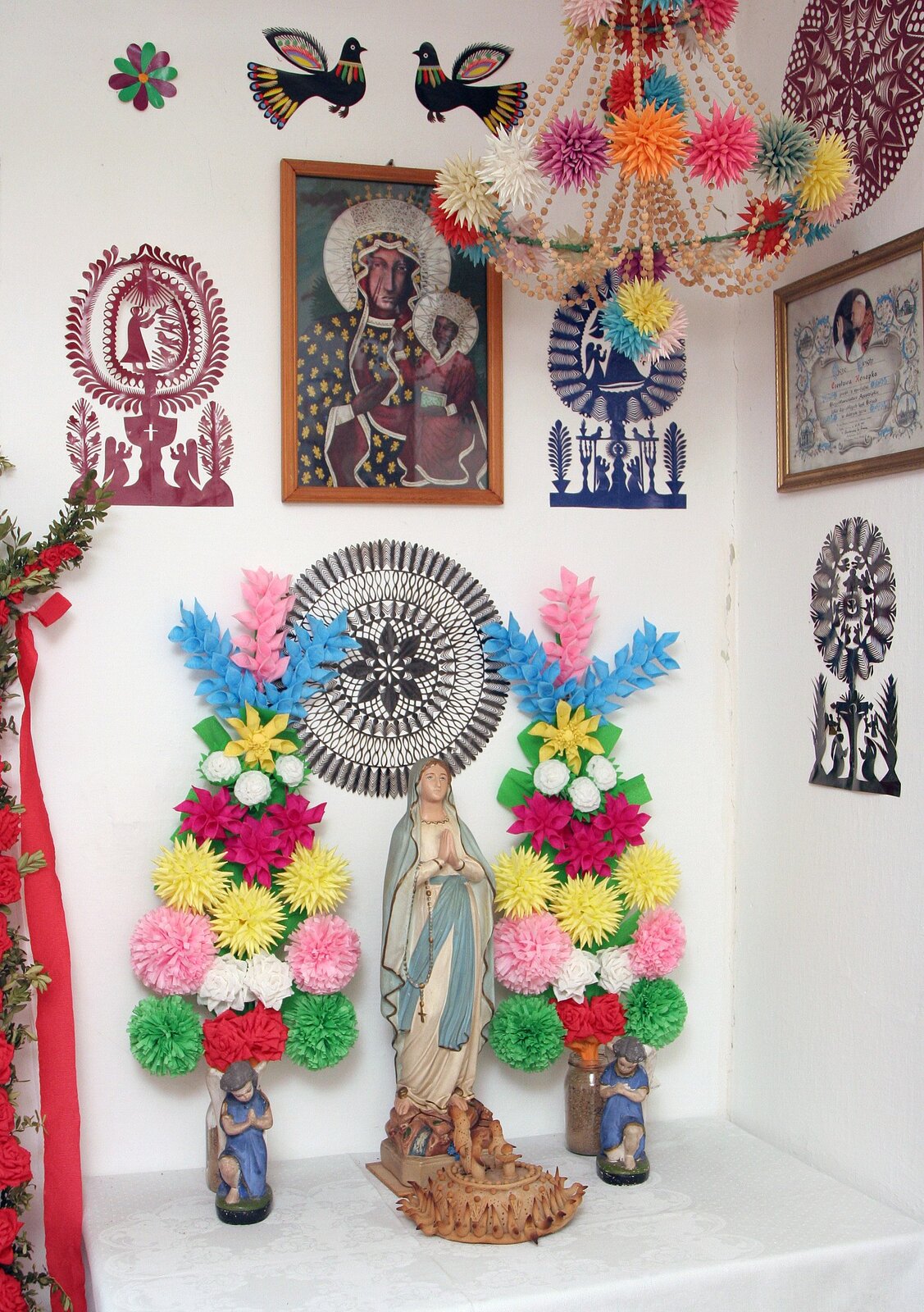 Fotografia kolorowych wycinanek pozawieszanych na ścianie obok świętych obrazów.