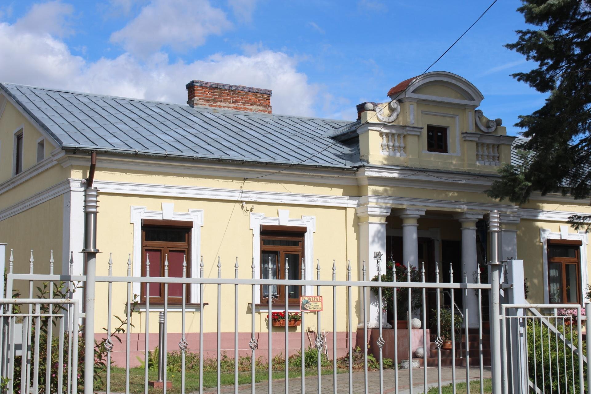 Zdjęcie przedstawia parterowy budynek ze skośnym, szarym dachem. Elewacja budynku jest w kolorze żółtym z elementami białymi. Okna mają brązowe ramy. Wejście do budynku jest bogato zdobione - znajdują się przy nim kolumny oraz część górująca nad dachem, wieńcząca wejście. Budynek znajduje się za szarą bramą. Niebo jest błękitne, częściowo przysłonięte przez białe chmury. Zdjęcie zostało wykonane w świetle dziennym.