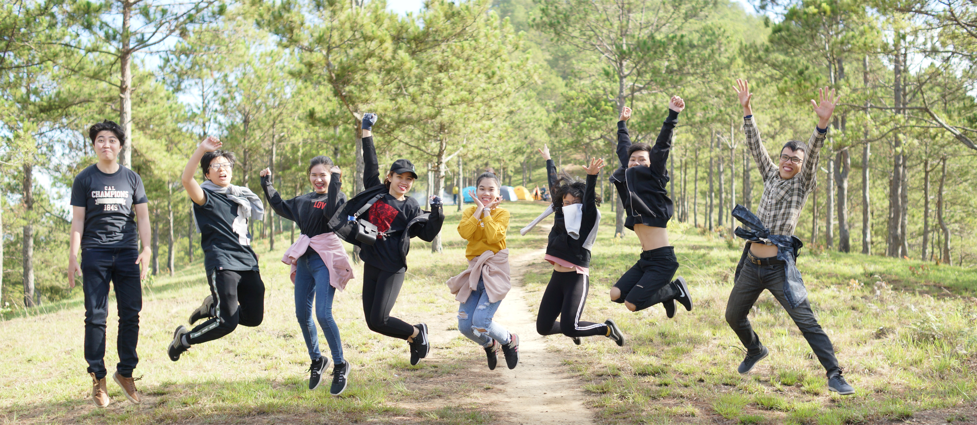 Zdjęcie przedstawia ośmioro nastolatków. Młodzi ludzie podskakują w geście radości. Ręce mają uniesione nad głową. Zdjęcie zostało wykonane na ścieżce w lesie.