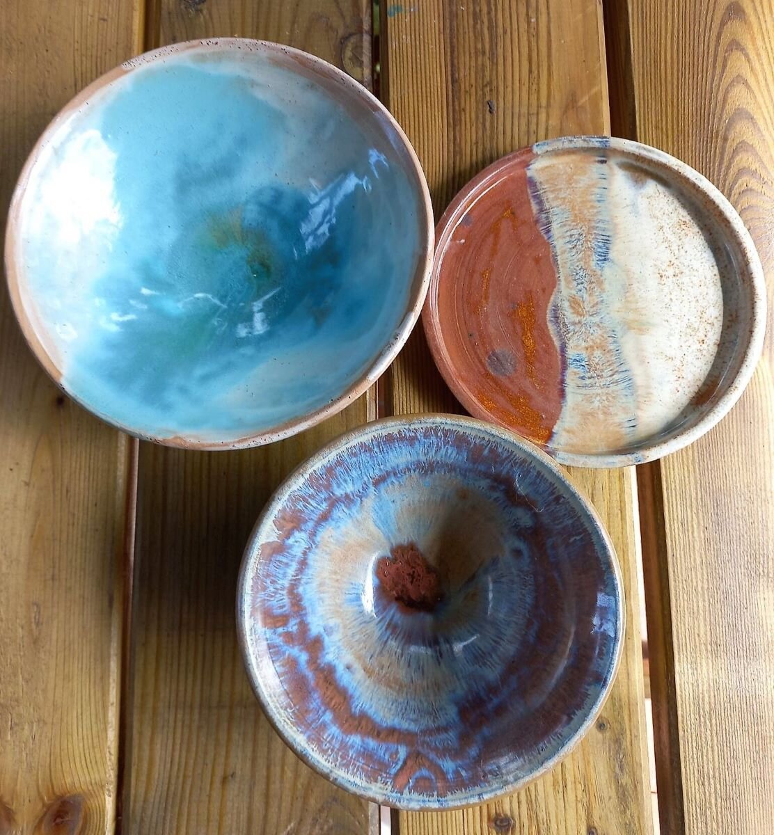 Zdjęcie przedstawia trzy naczynia ceramiczne pokryte zróżnicowanym szkliwem. Są to dwie miseczki i spodek, stojące na drewnianym blacie stołu.