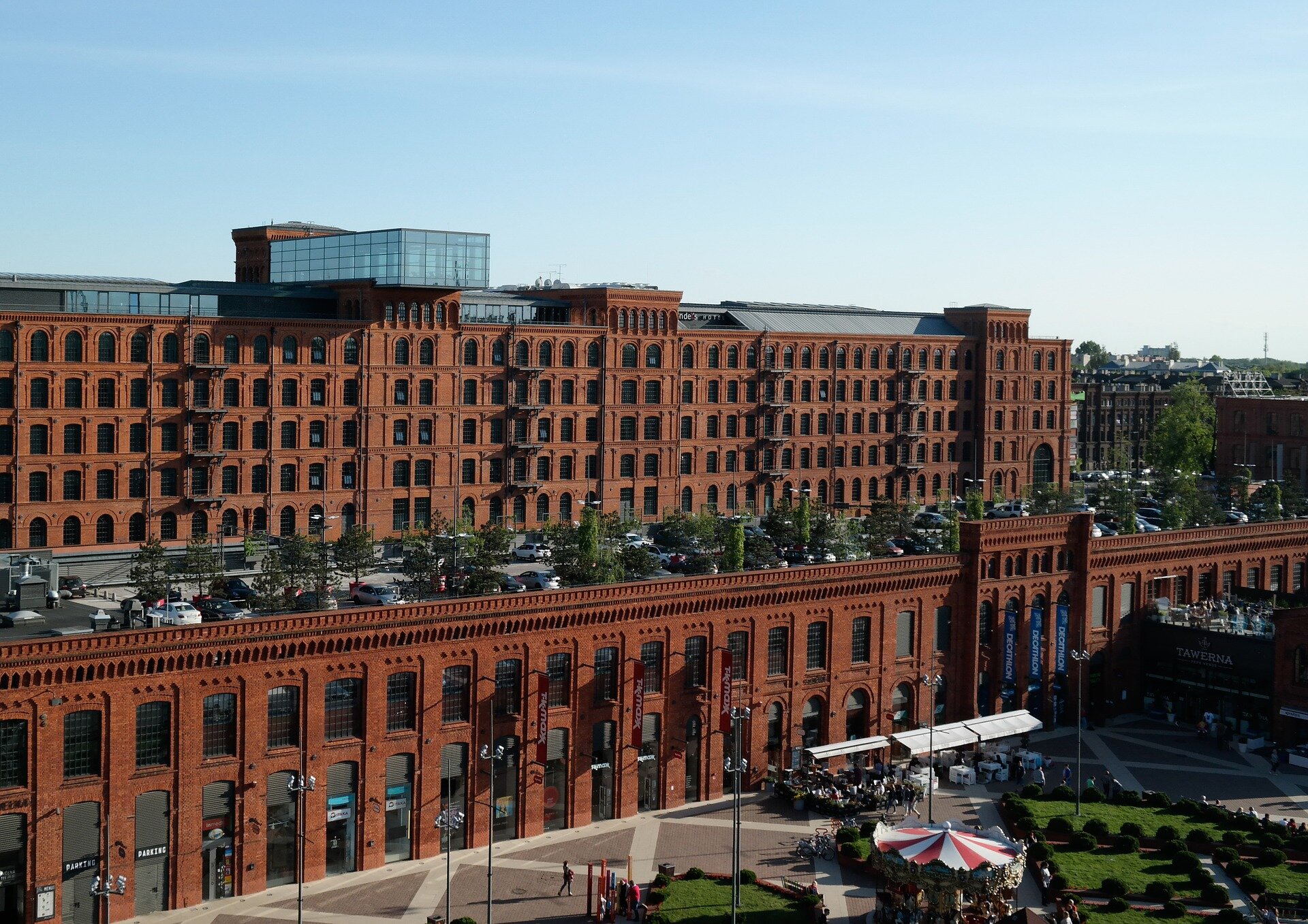 Zdjęcie przedstawia budynek z zewnątrz. Jest wykonany z czerwonej, nieotynkowanej cegły. Ma rzędy okien. Na dachu niższej części obiektu zaprojektowano parking.