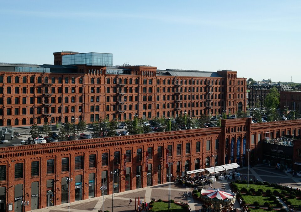 Zdjęcie przedstawia budynek z zewnątrz. Jest wykonany z czerwonej, nieotynkowanej cegły. Ma rzędy okien. Na dachu niższej części obiektu zaprojektowano parking. 